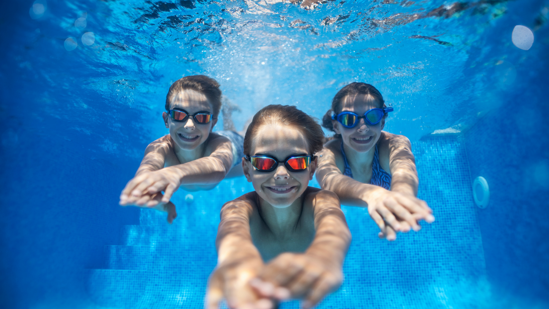 Three children swimming underwater in a blue pool, wearing goggles and smiling at the camera.