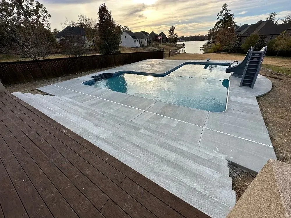 Pool with a slide, surrounded by a concrete patio and a wooden deck, near a lake.