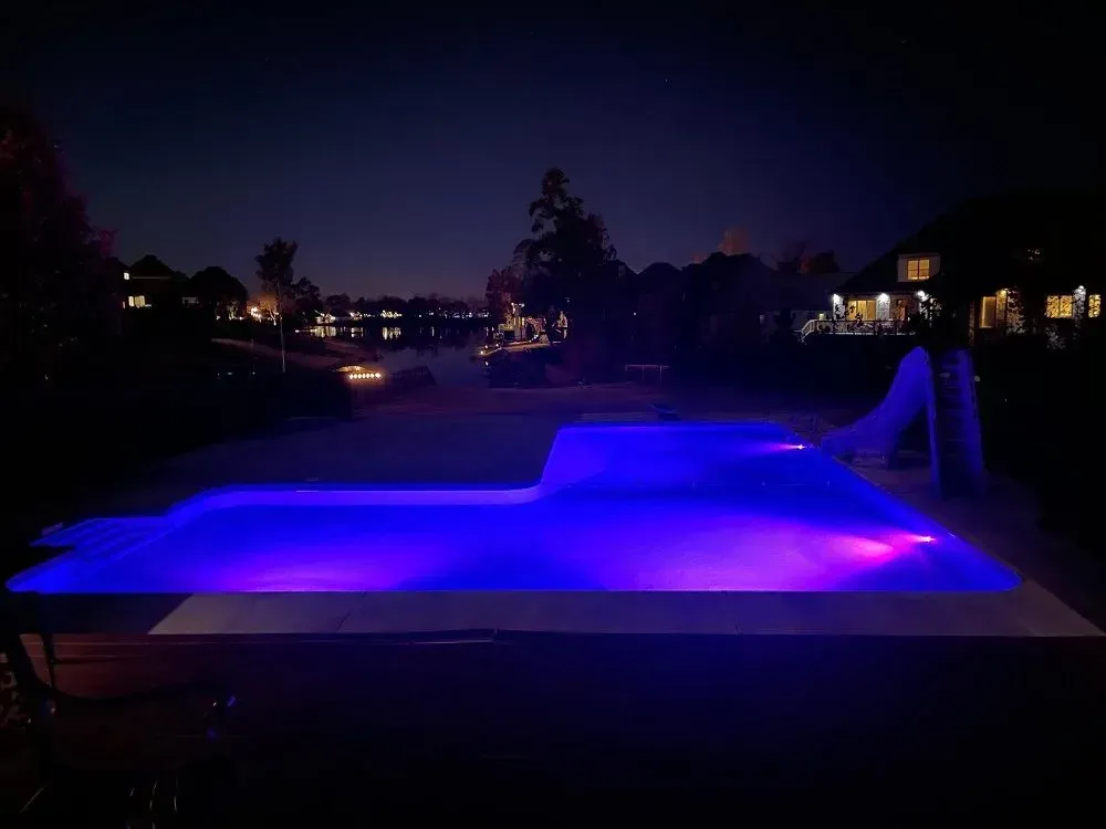 Nighttime view of a pool with blue and pink lights. Trees and houses are visible in the background.