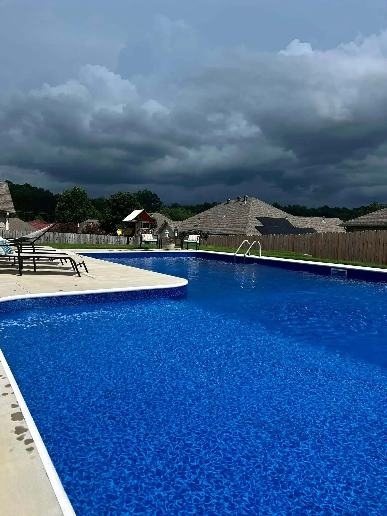 Swimming pool on a cloudy day, surrounded by homes and a playground. Blue water reflects sky.