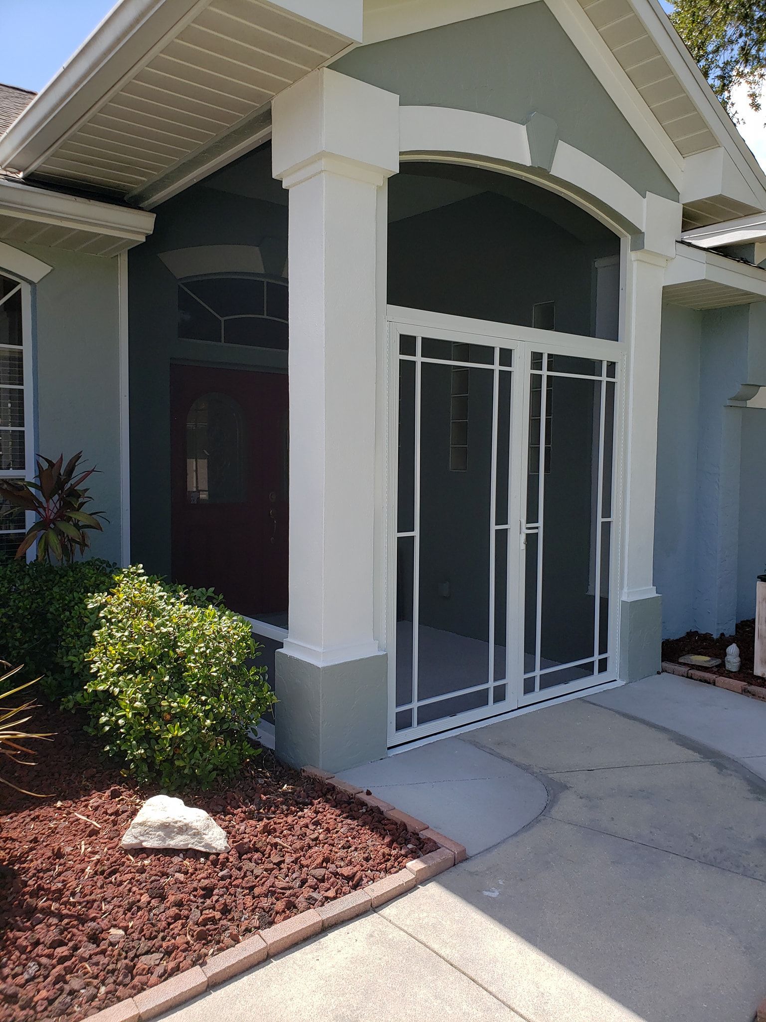 Screened-in porch with white metal doors and columns. Entryway of a house with light blue siding, red front door.