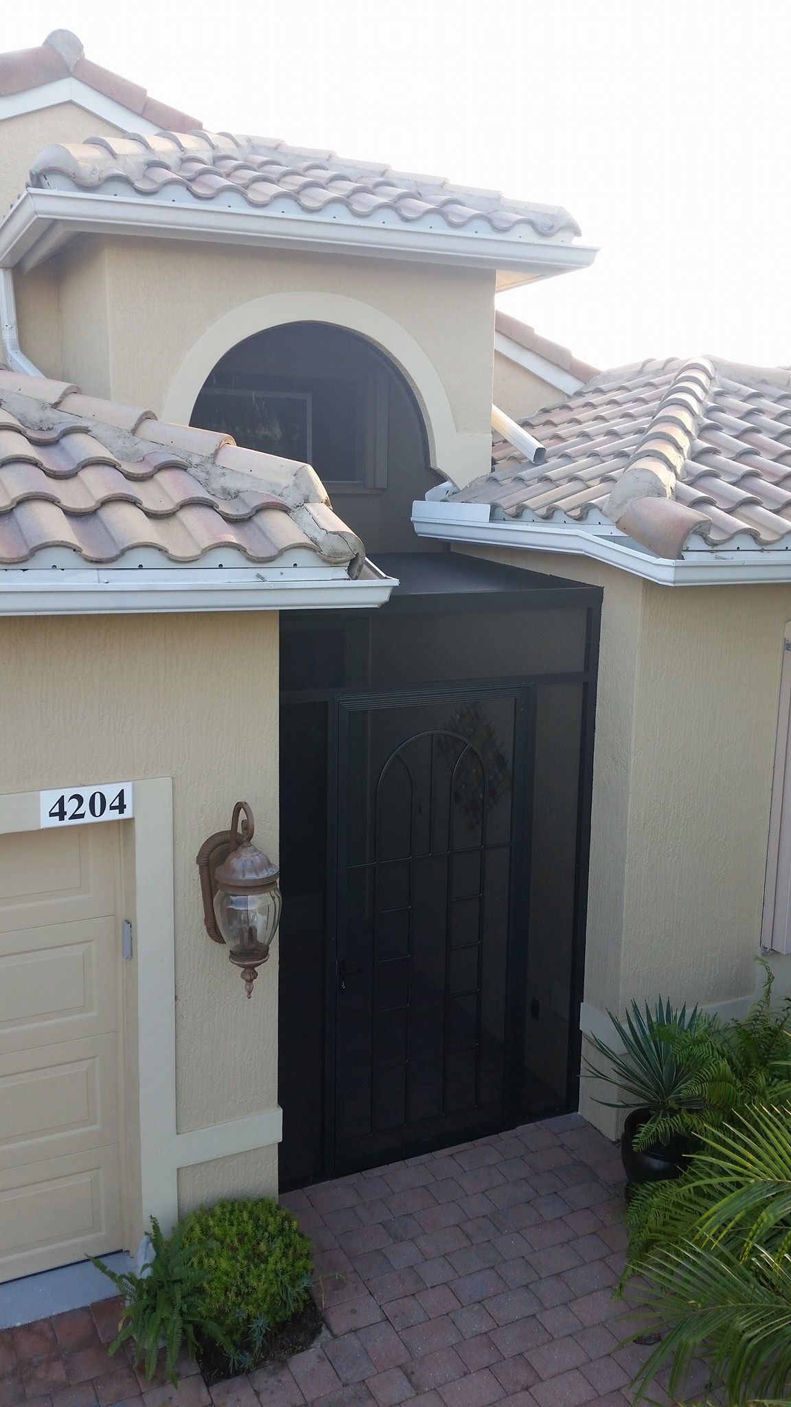 Black gate in front of a beige house with tiled roof and archway. A house number is on the left.