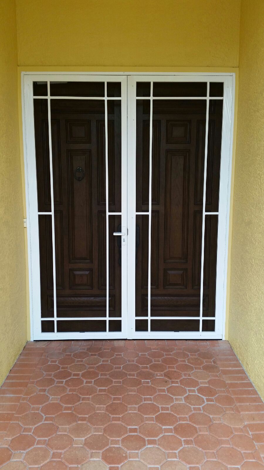 Double brown doors with white security bars on a yellow wall with red-brown tile flooring.