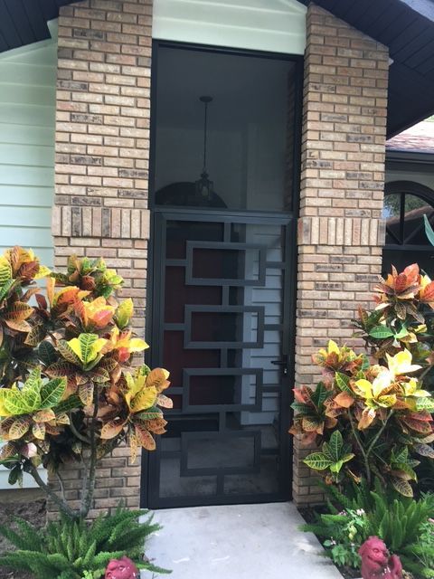 Black screen door with geometric design, brick columns, and colorful plants flanking the entrance.