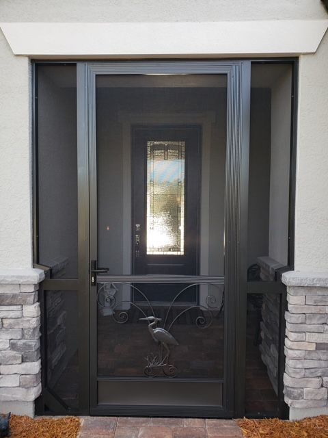 Dark screen door with heron detail, leading to a front door with glass, set between stone columns.