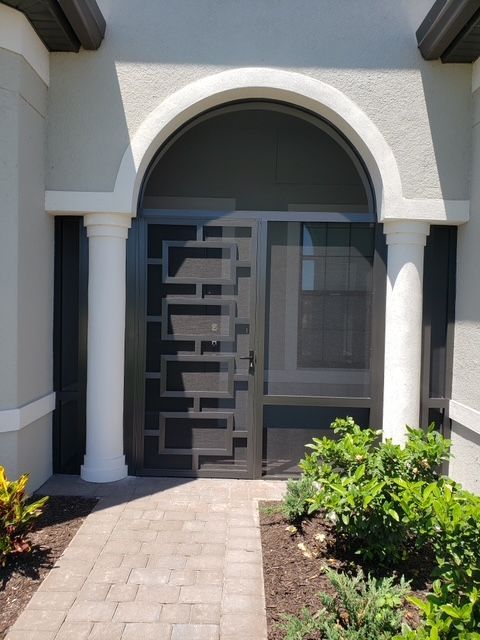 A black metal front door with geometric design, arched window above, flanked by white columns, on a brick path.