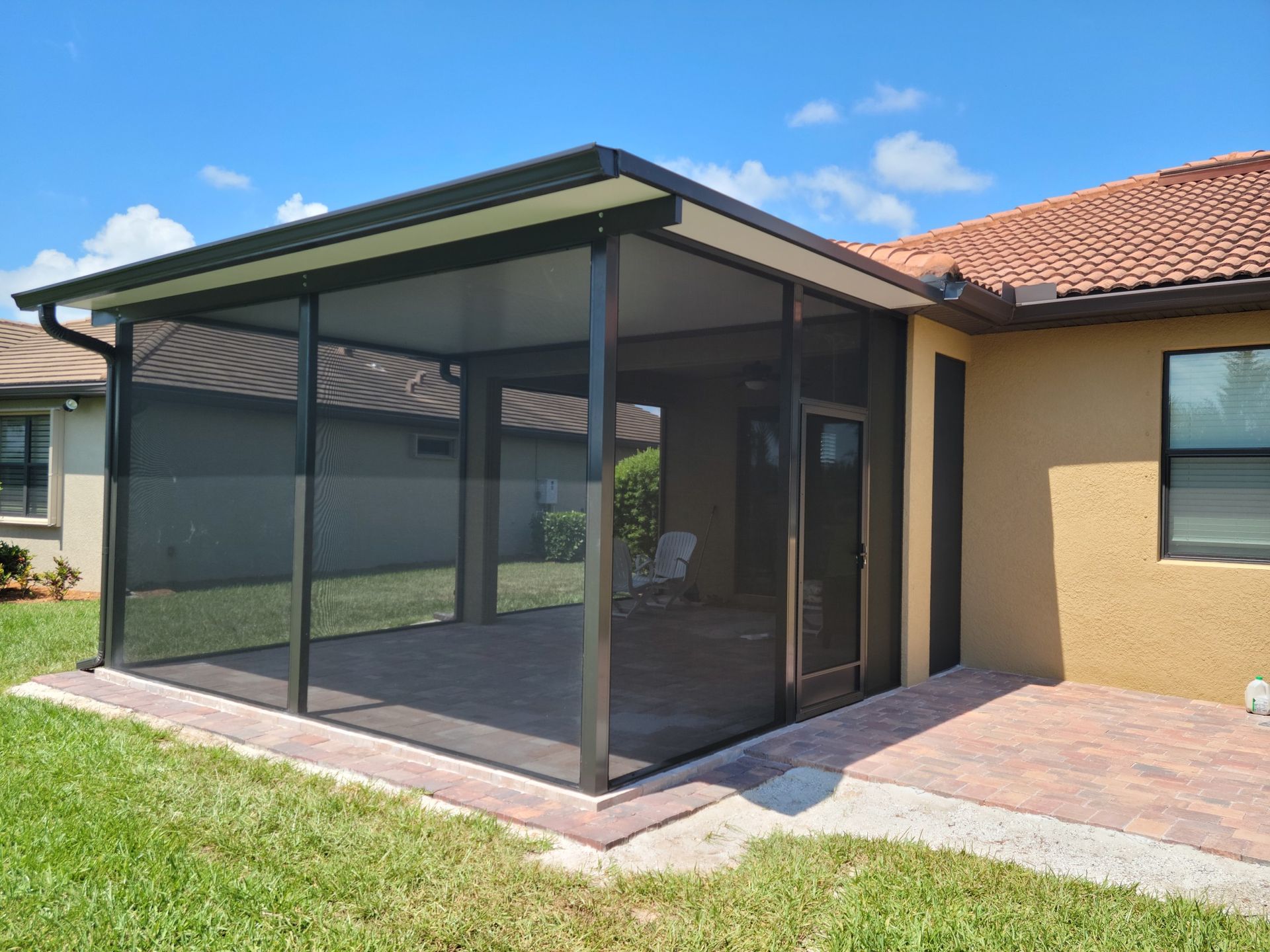 Screened-in patio extension attached to a stucco house with a tiled roof. Black frames and screening. Outdoors, sunny.