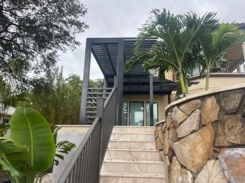 Stone steps lead up to a building with a dark pergola, palm trees, and bamboo in the background under a cloudy sky.