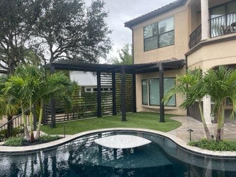 Backyard with pool and dark pergola near a house. Green grass, palm trees, and overcast sky.