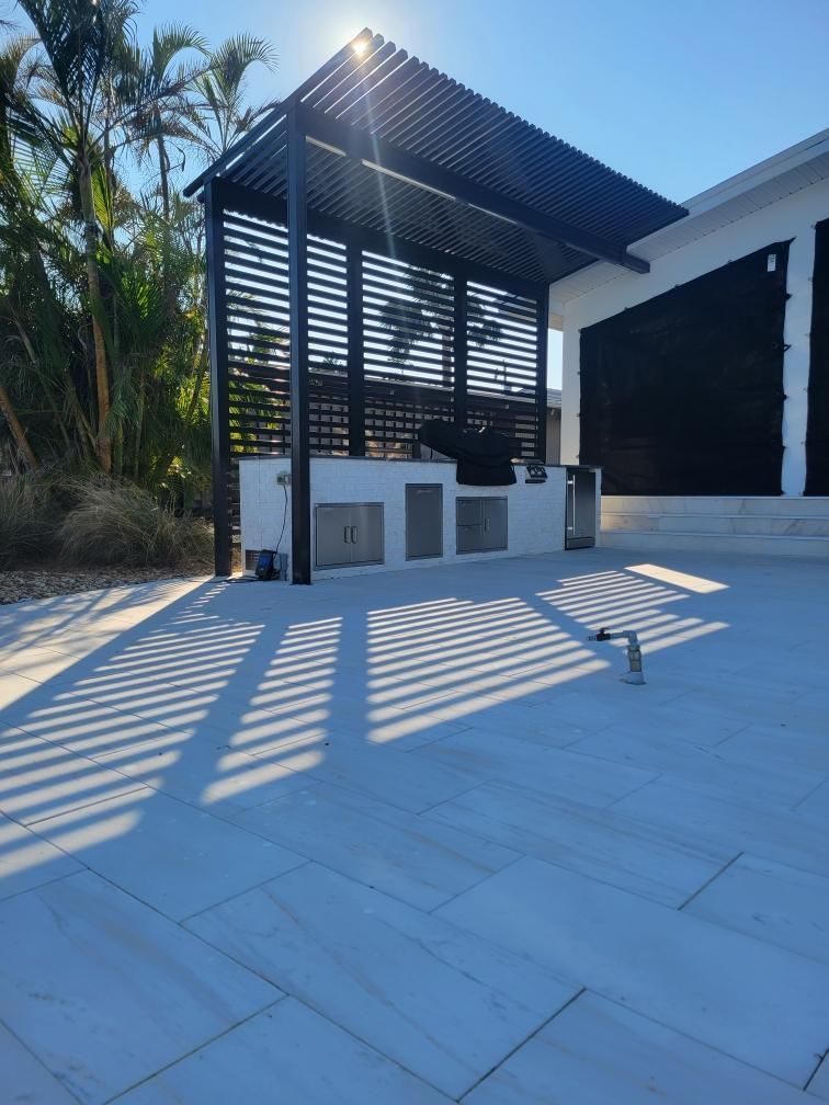 Outdoor kitchen with black pergola and white countertops on a stone patio, casting striped shadows.