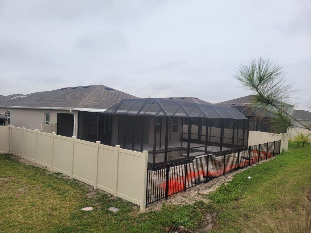 Fenced backyard with a screened pool enclosure and adjacent house under a cloudy sky.