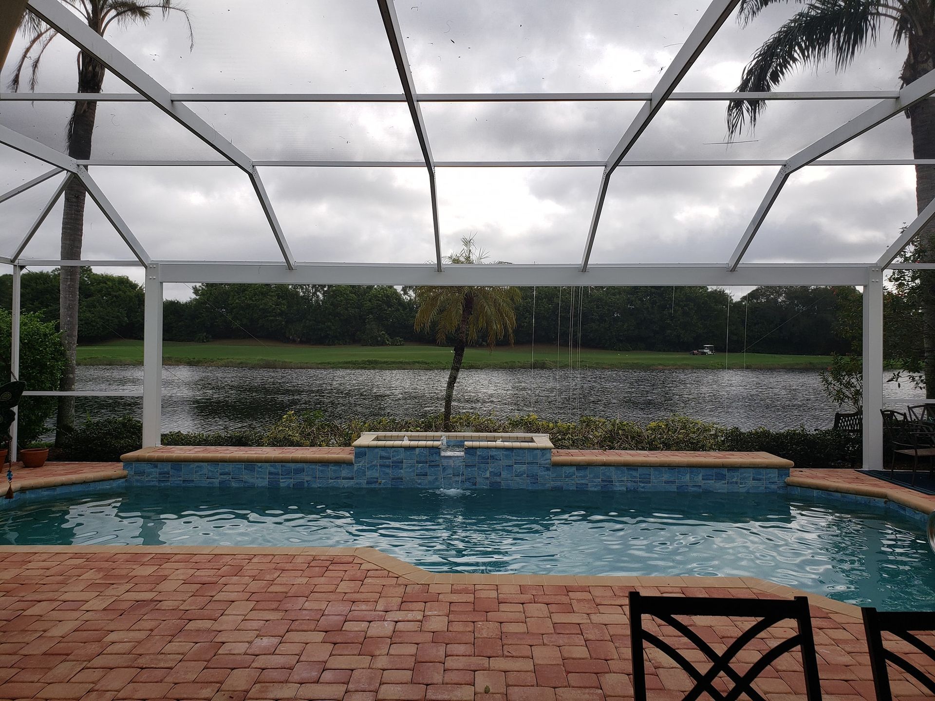 Pool with brick patio, screened enclosure, overlooking a lake under a cloudy sky.