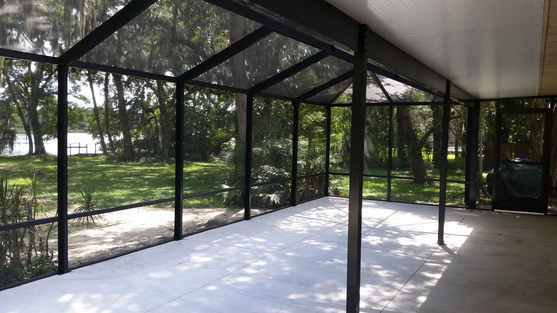 Screened-in porch with black framing and light gray concrete floor, overlooking a grassy yard with trees and a lake.