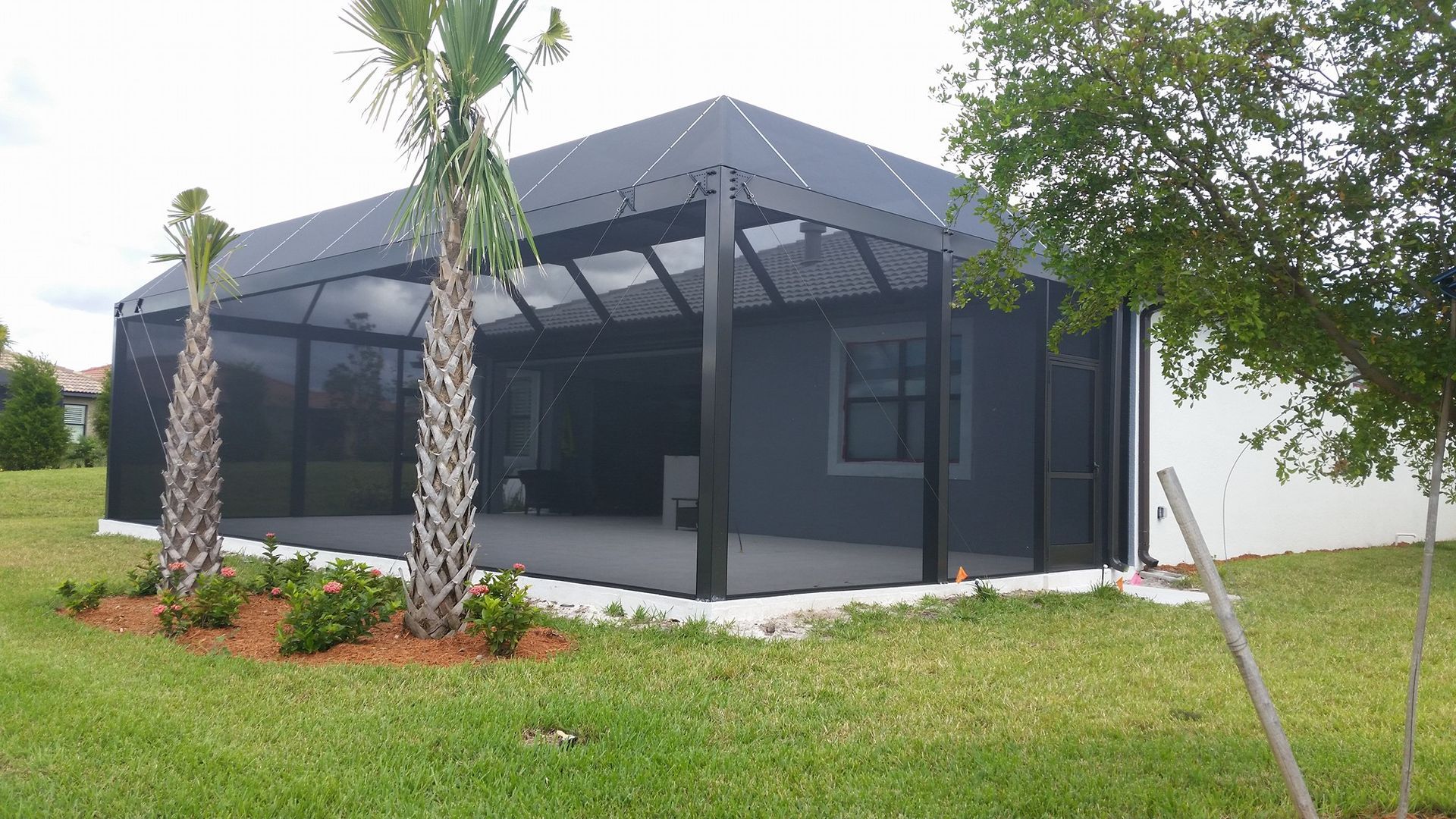 Screened patio attached to a white house, black frame, gray floor. Lawn in foreground, palm trees.
