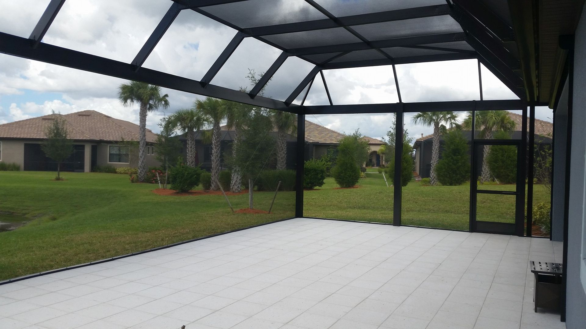 Screened-in porch with white deck floor, black frame, and view of a green yard and houses under a cloudy sky.