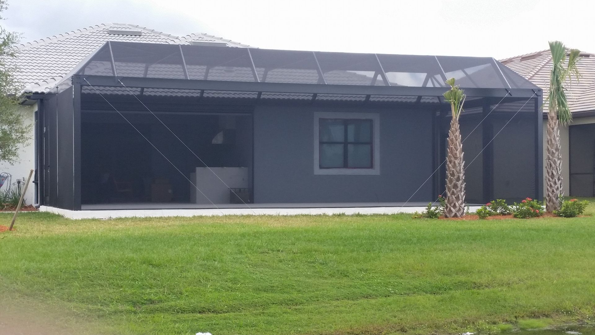 Screened patio attached to a gray house, with a dark screen enclosure and a green lawn.
