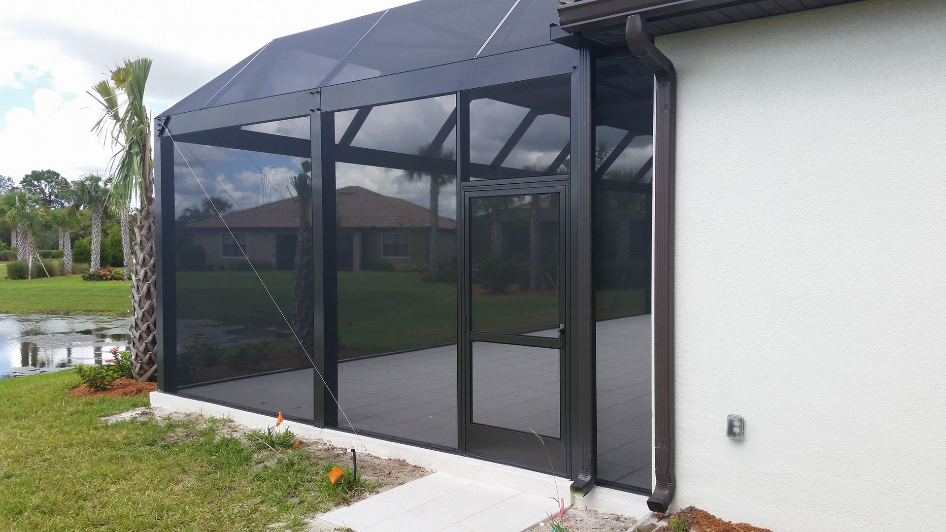 Screened patio enclosure with black frame, next to a white house, overlooking a grassy yard and pond.
