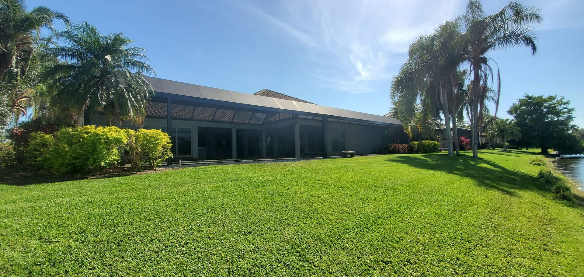 Lush green lawn in front of a long, dark-roofed building and palm trees under a blue sky.
