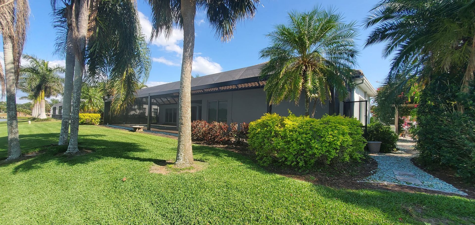 A house with a dark roof surrounded by green grass and palm trees under a blue sky.