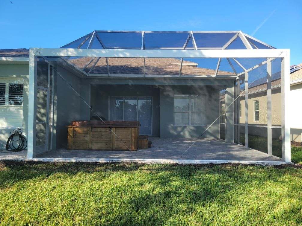 Screened-in patio with a hot tub, on a paved area, attached to a house with a blue sky in the background.