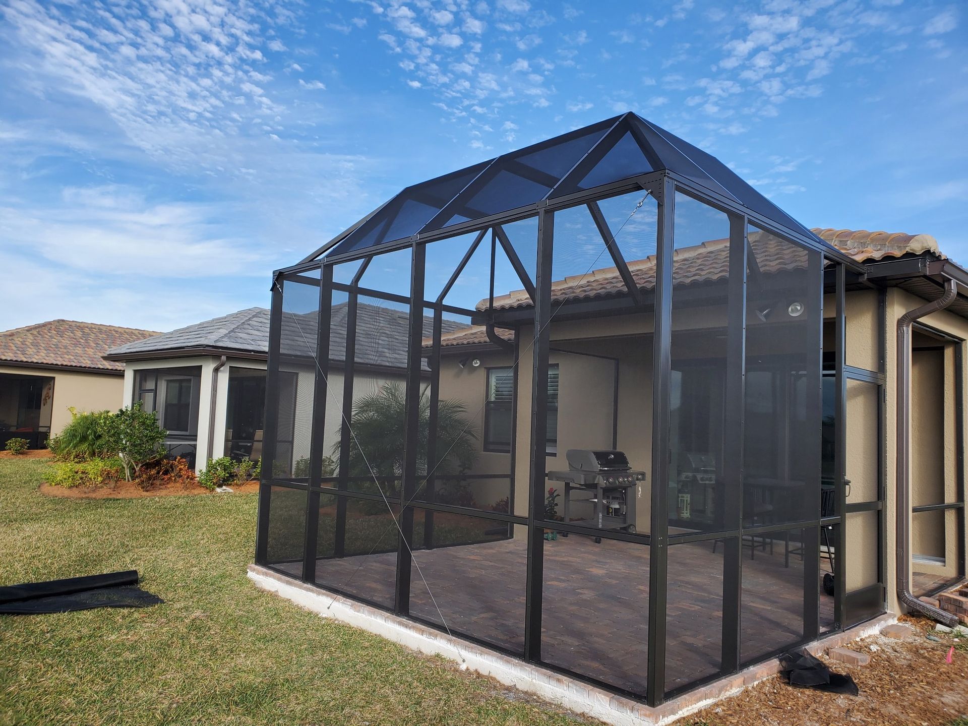 Screened-in patio with black framing and roof, attached to a light-colored house, in a grassy yard under a blue sky.