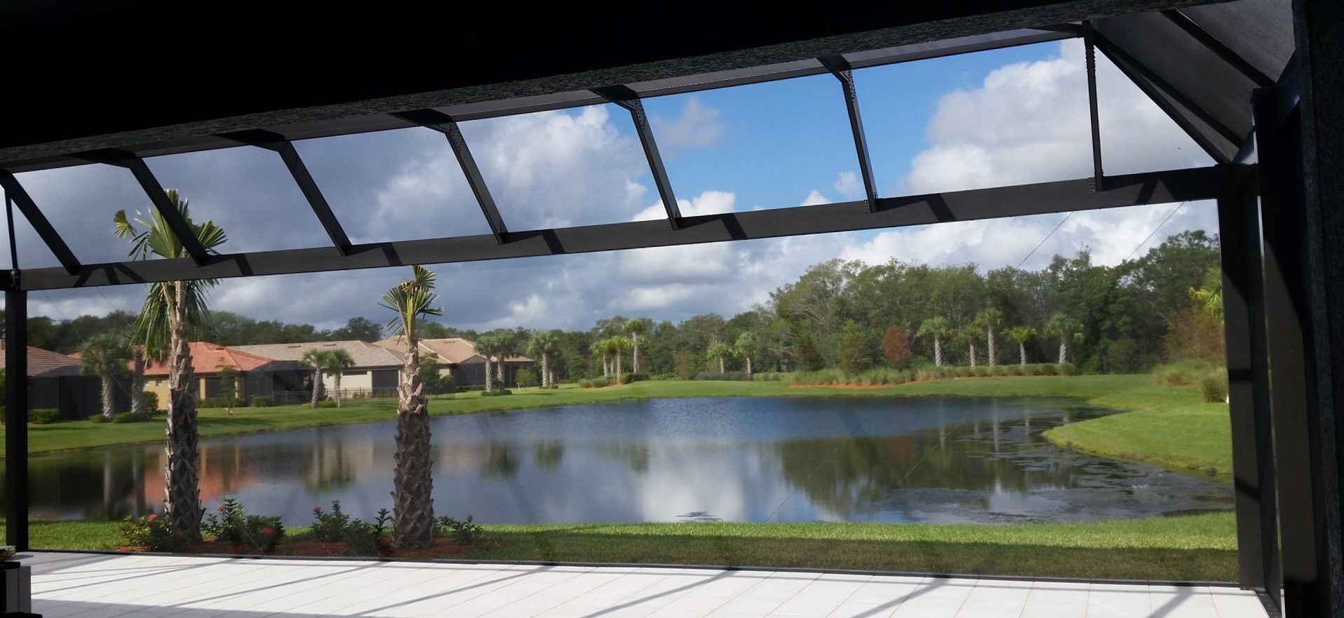 View of a pond from a screened patio. Blue sky and clouds reflected in the water. Houses and trees in the background.