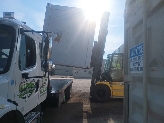 A white truck and a yellow forklift are lifting a large white box outdoors on a sunny day.
