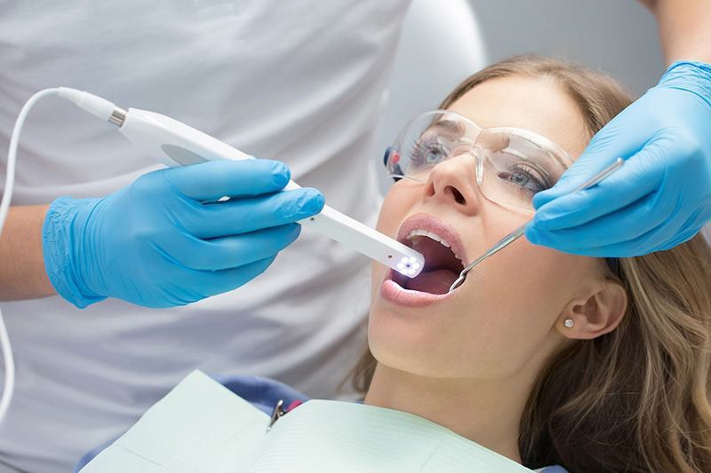 Dentist using a dental scanner on a patient's open mouth; blue gloves, protective eyewear.