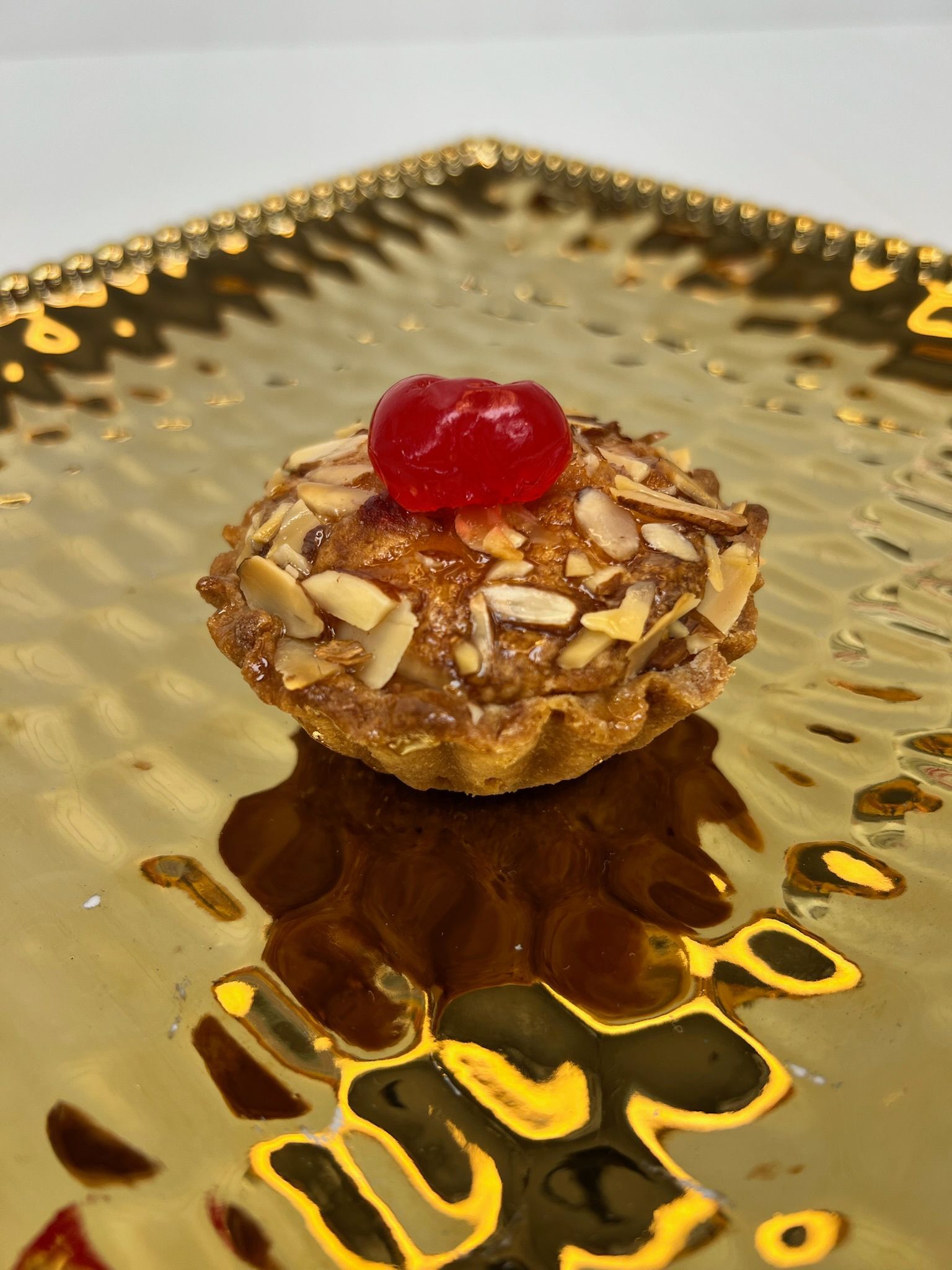 Small pastry with sliced almonds and a cherry on a gold, textured plate.
