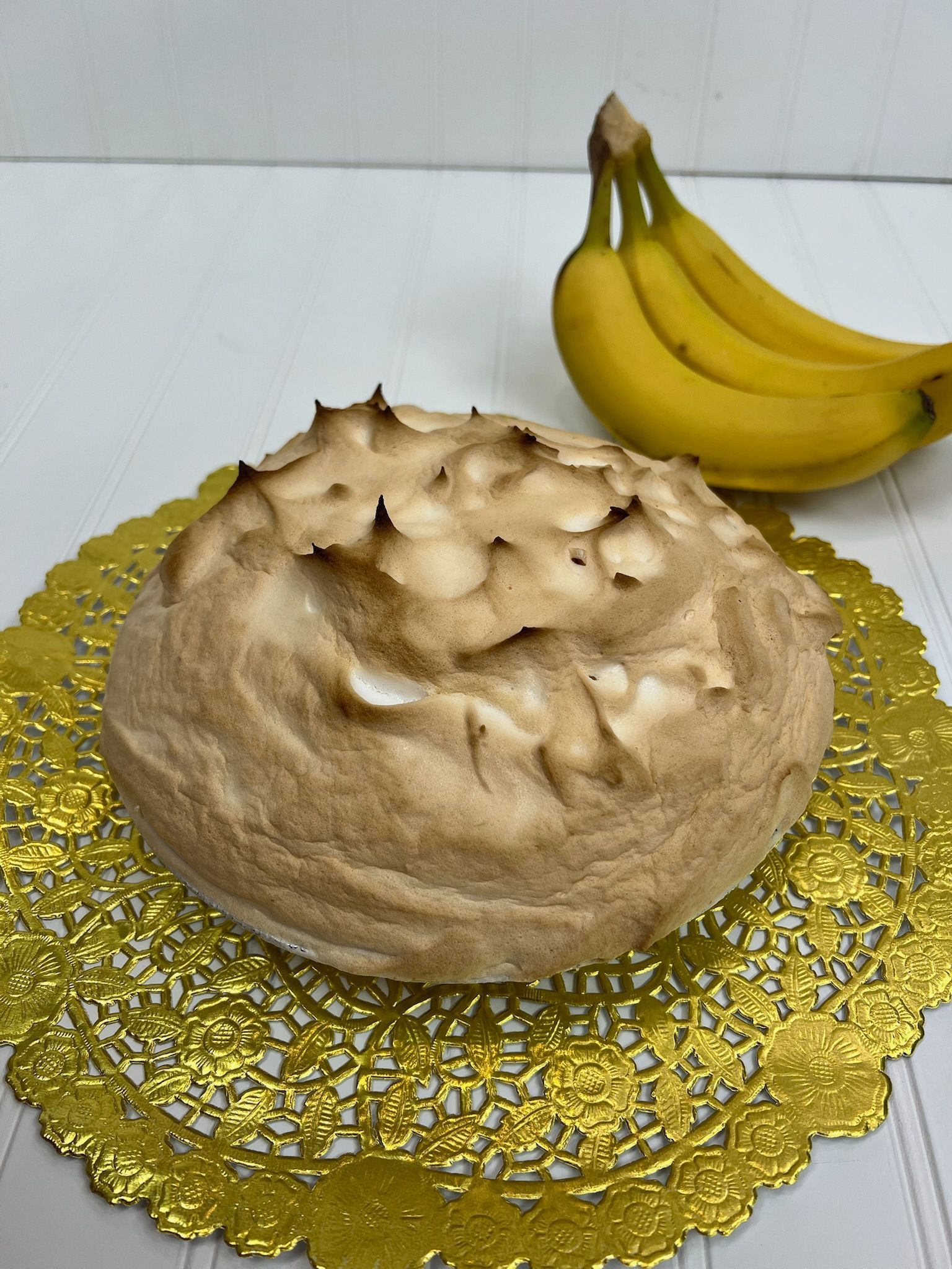 Toasted meringue dessert on a gold doily, with bananas in the background.
