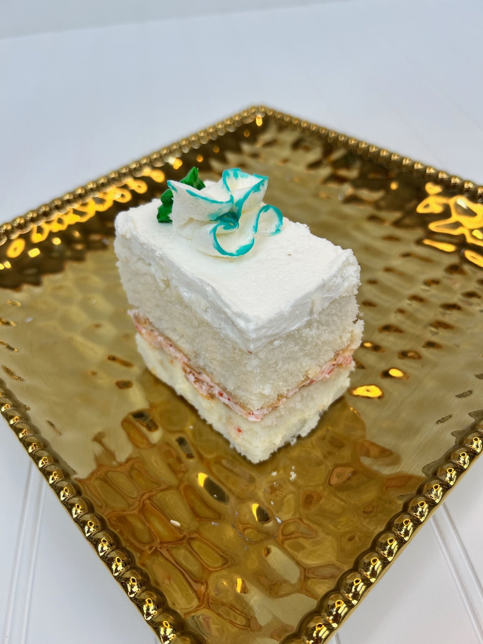 Cake slice with white frosting and teal flower on a gold square plate.