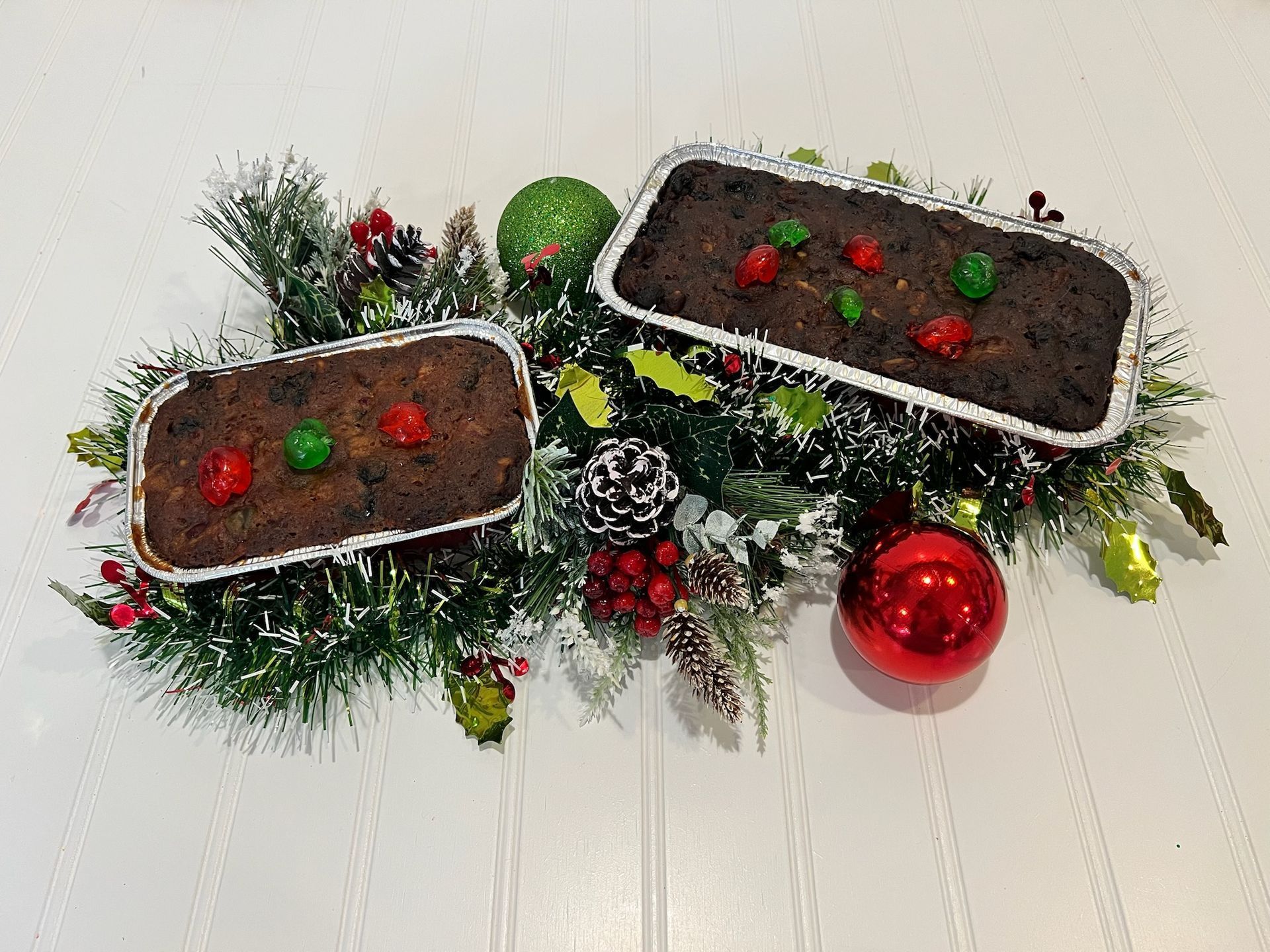 Two fruitcakes decorated with cherries, atop Christmas greenery, with ornaments.