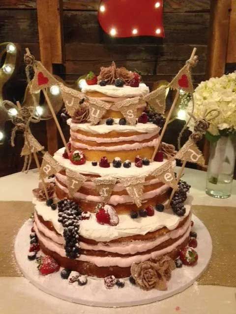 Rustic three-tier wedding cake with "Just Married" banner, berries, flowers, and fairy lights.