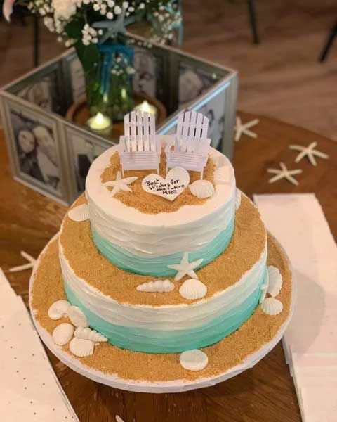 Two-tiered beach-themed cake with blue and white frosting, sand details, seashells, and Adirondack chair toppers.