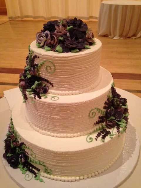 Three-tier white cake decorated with dark purple and green floral accents, topped with flowers.