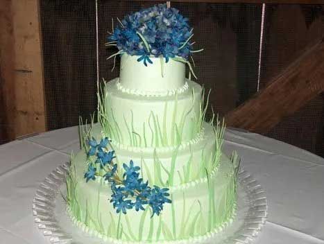 Three-tiered cake with light green frosting, blue flower decorations, and grass accents, on a white tablecloth.