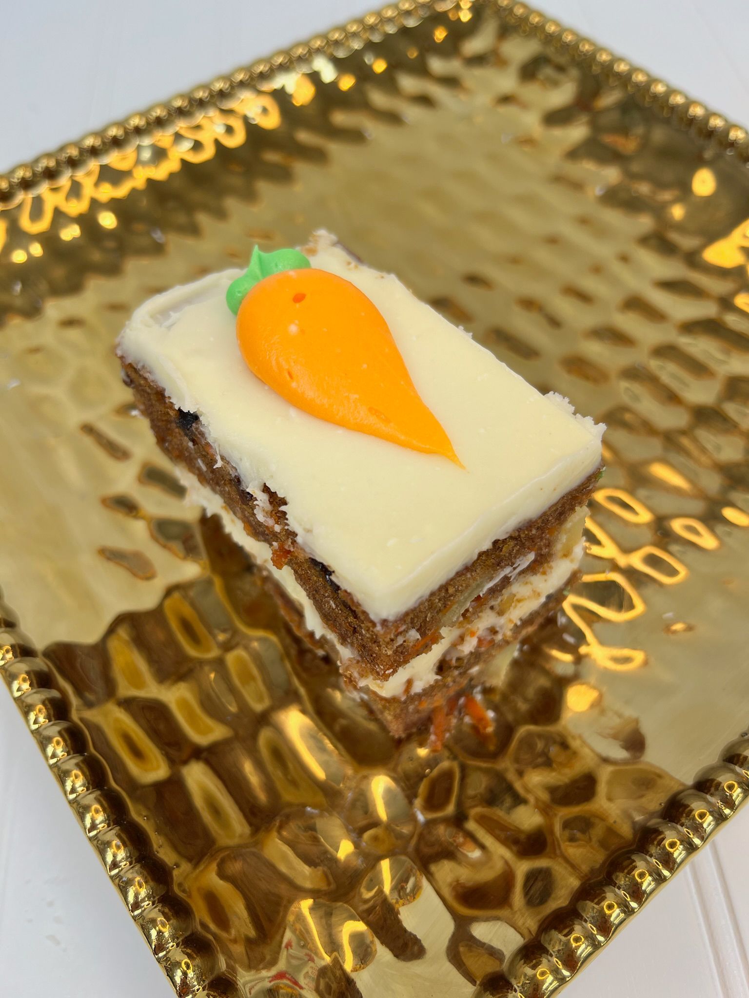Carrot cake square with white icing and an orange carrot decoration on a gold patterned plate.