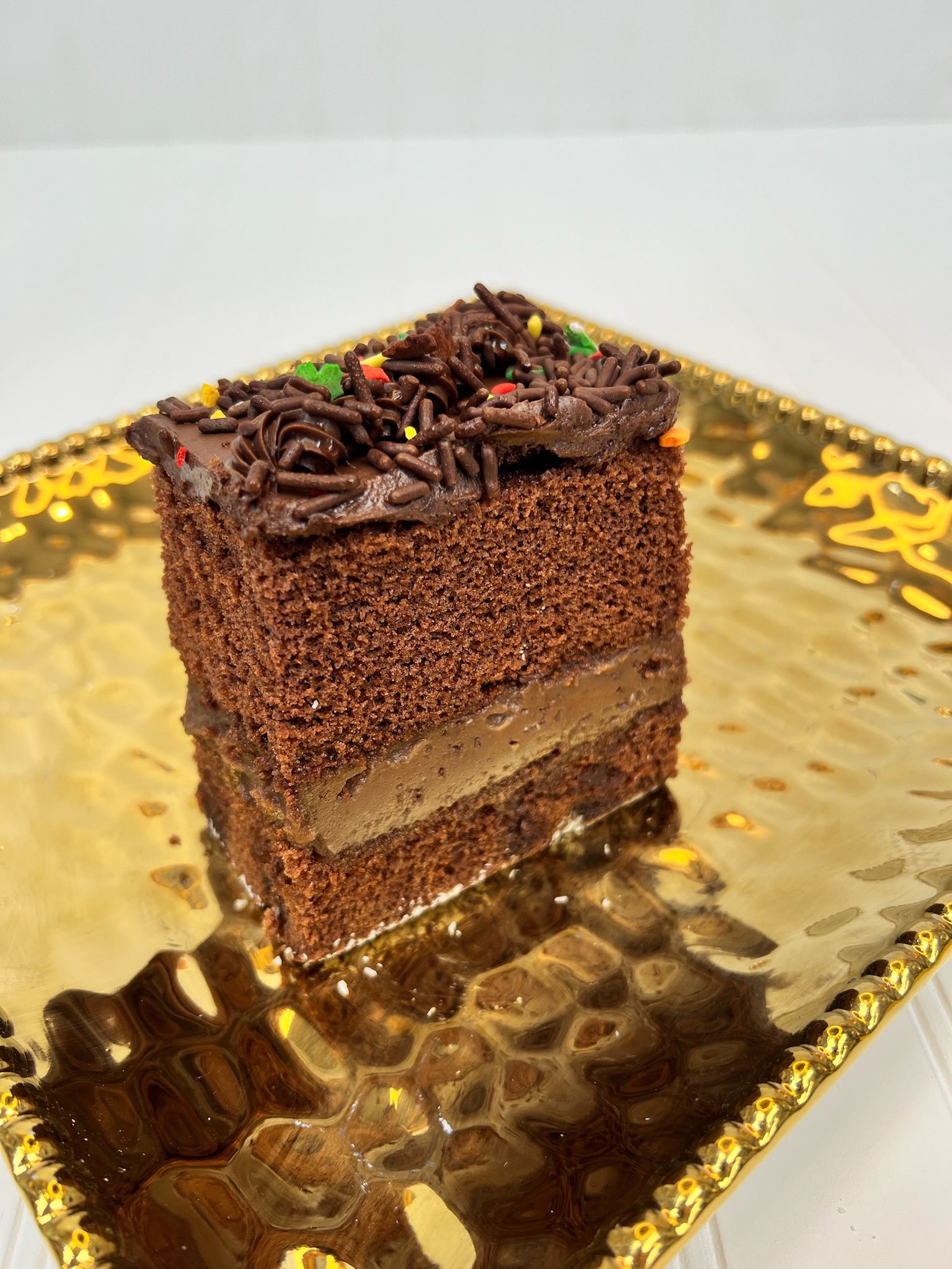 Chocolate cake slice on a gold platter, sprinkled with chocolate and rainbow sprinkles.