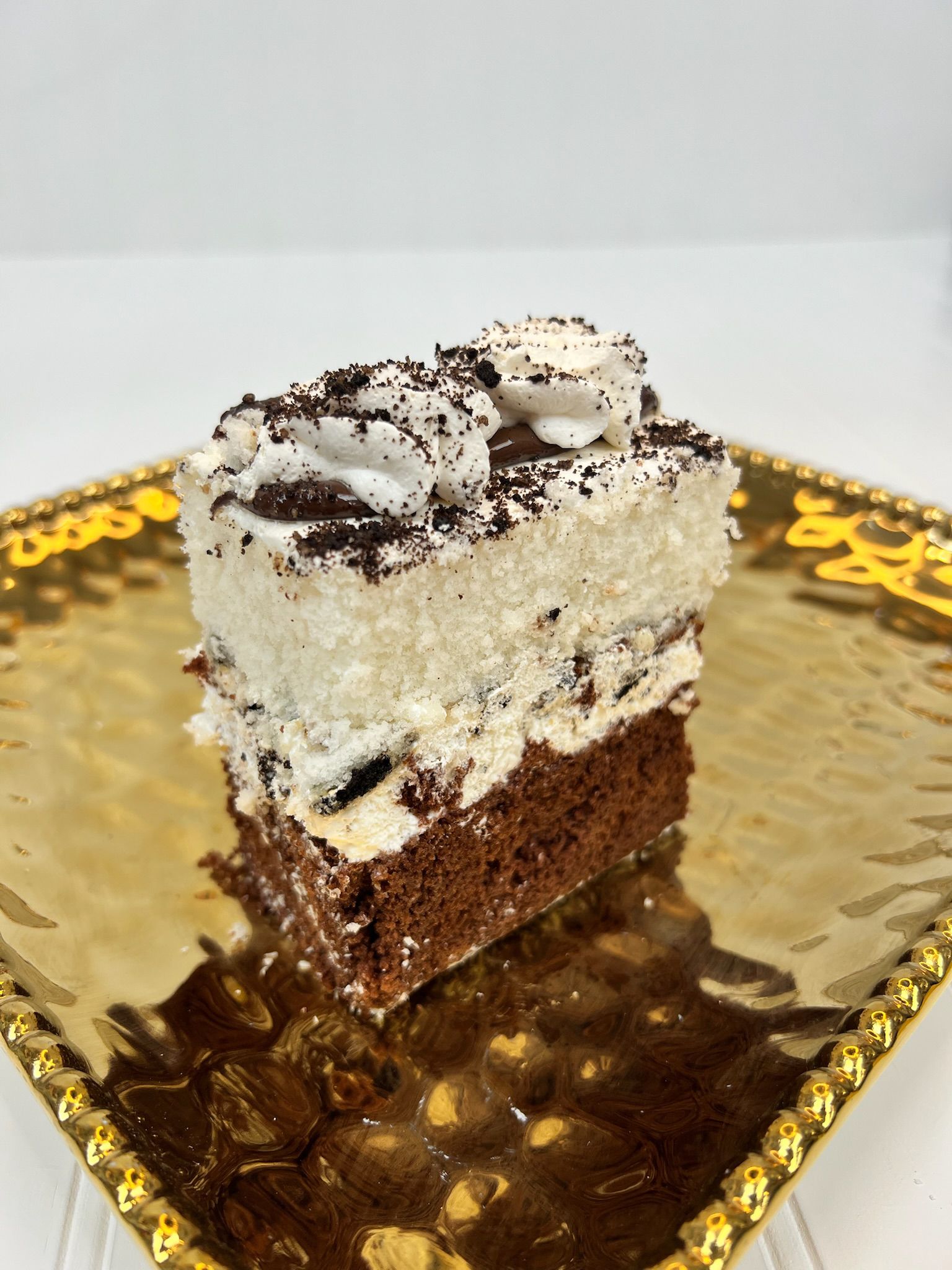 Chocolate cake slice on a gold-colored plate, topped with white frosting and dark sprinkles.