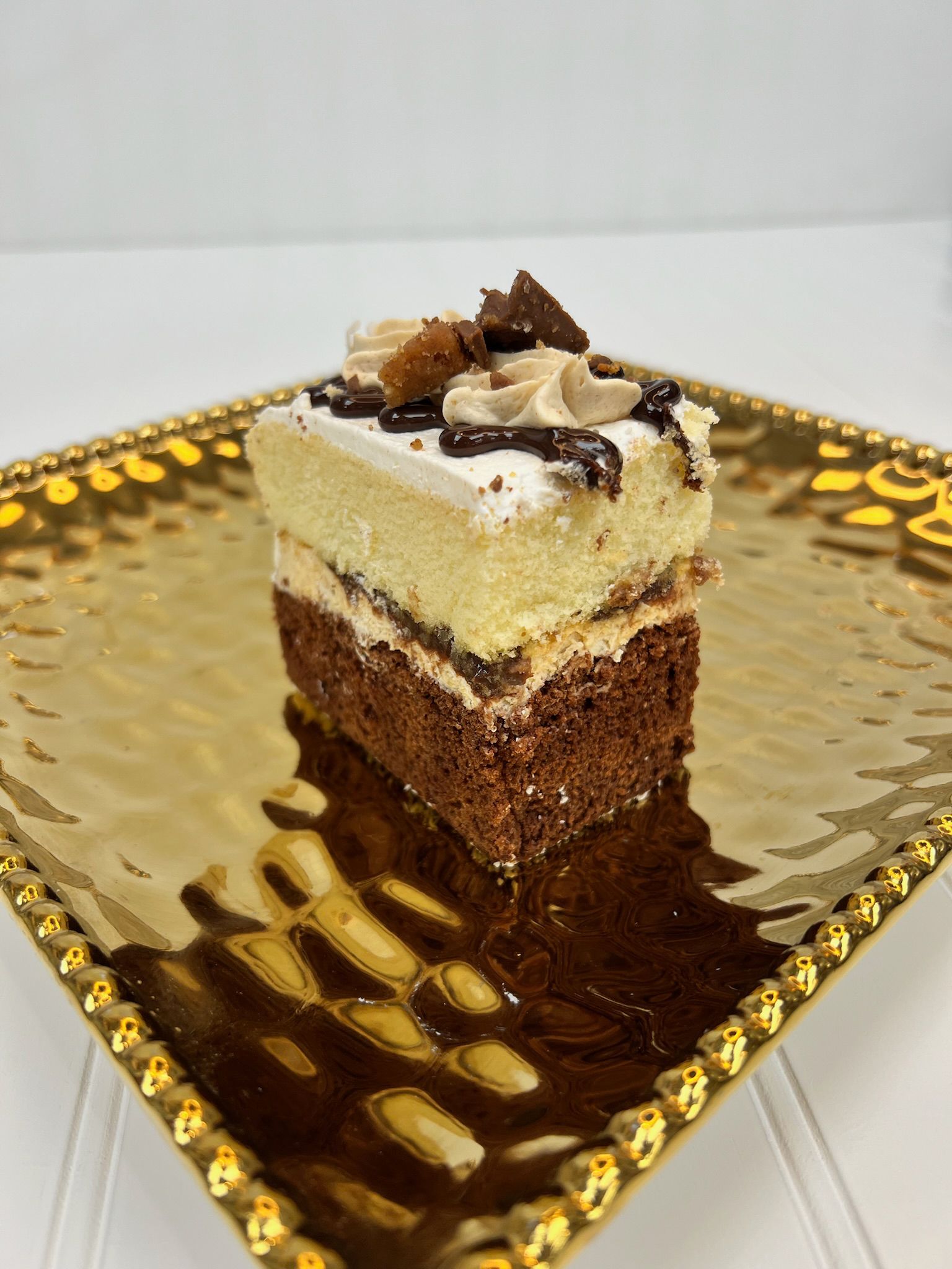 Cake slice on a gold, patterned plate with brown and white layers, topped with frosting and chocolate chunks.