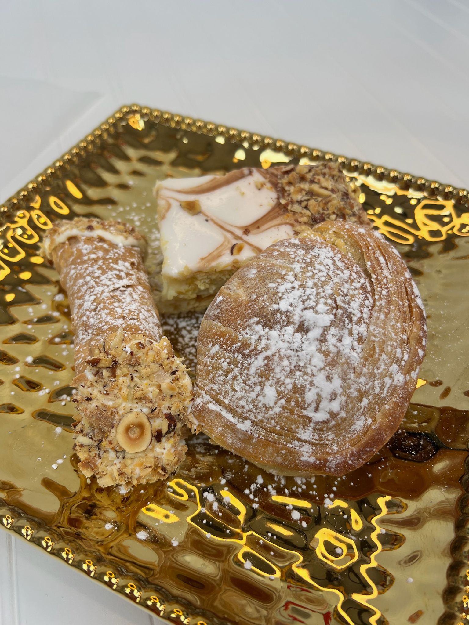 Pastries on a gold textured plate, dusted with powdered sugar. Includes a cannoli, cake, and croissant.