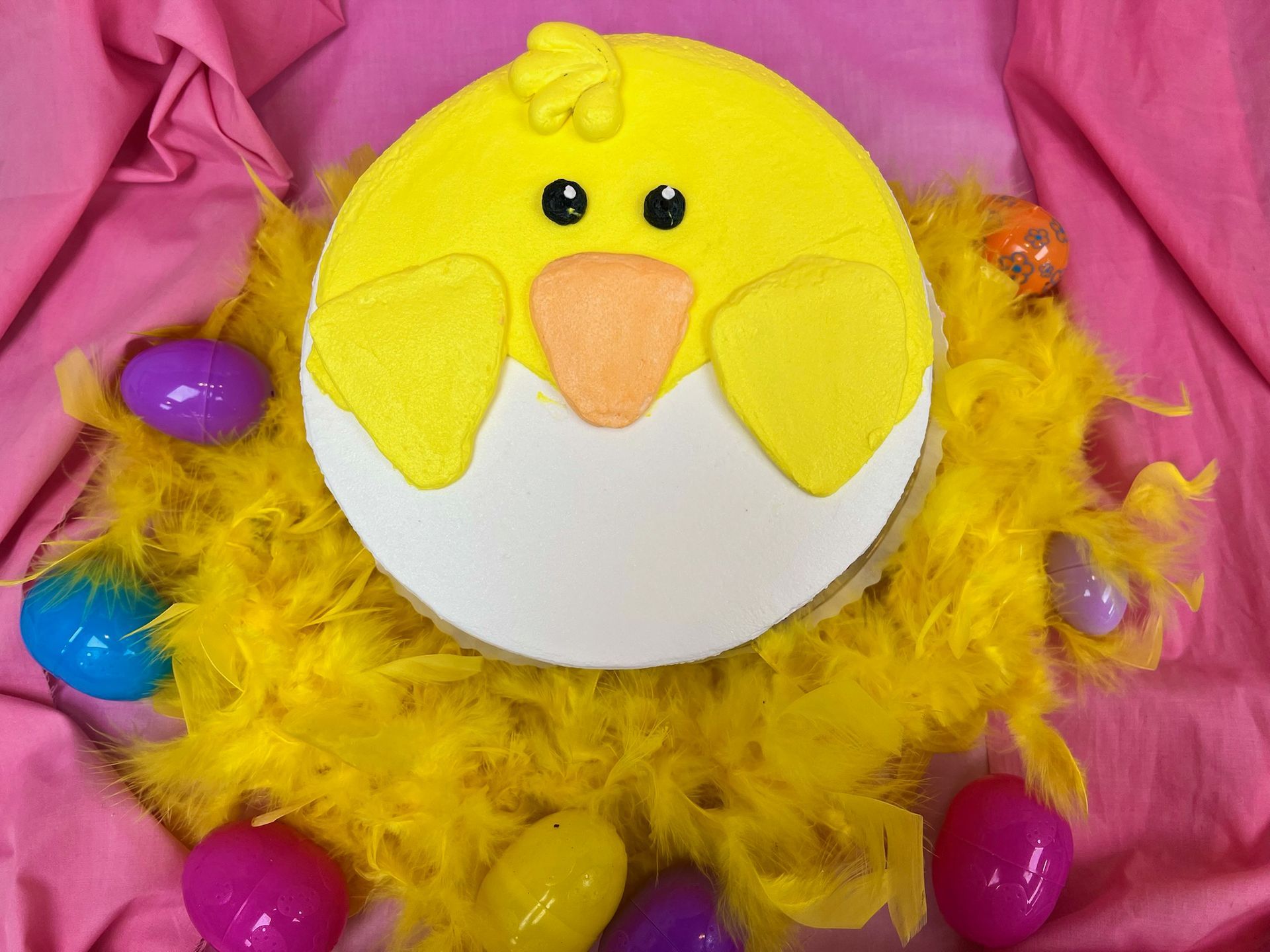 Yellow chick cake with an eggshell, surrounded by yellow feathers and Easter eggs.