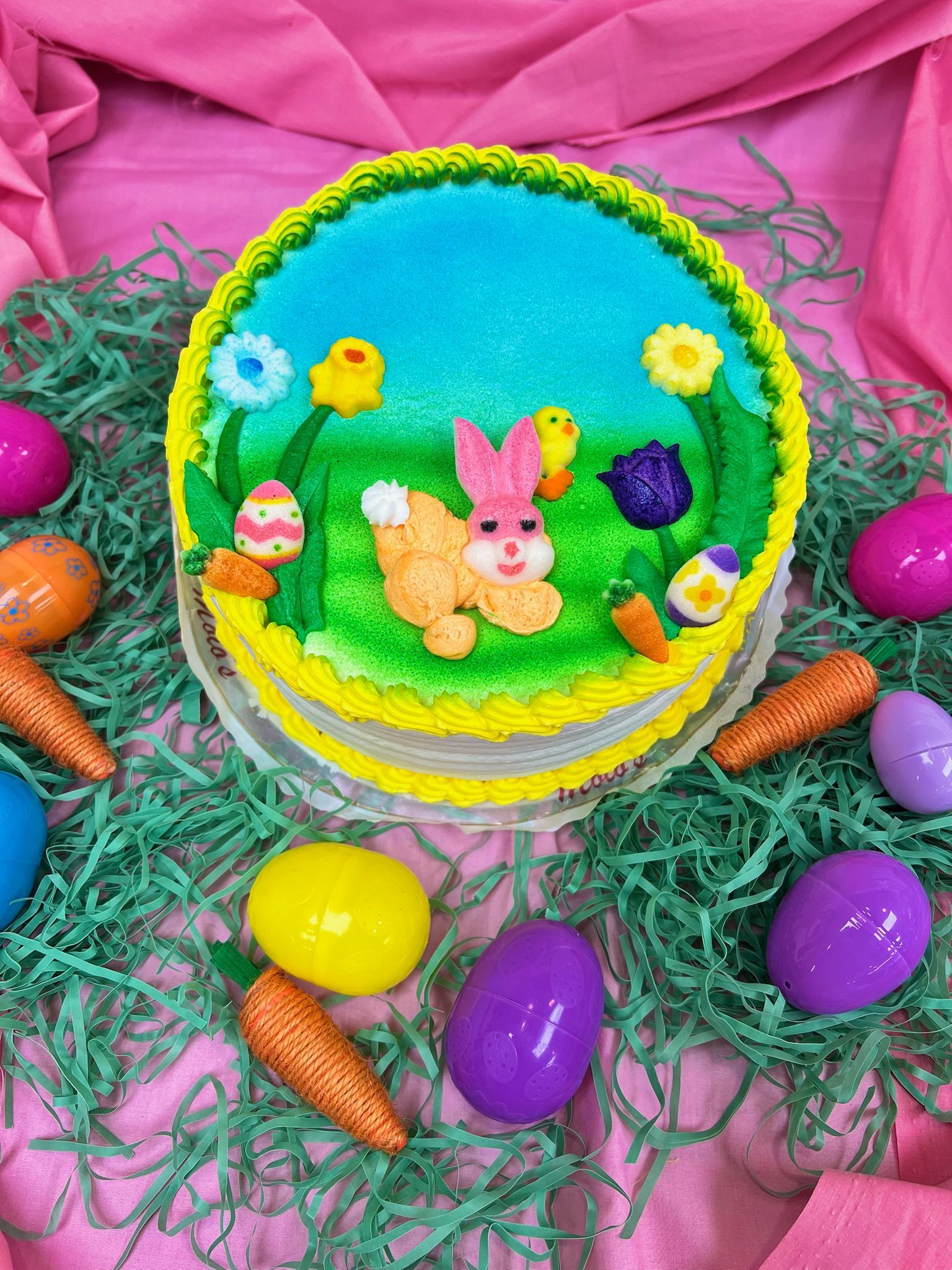Easter cake with bunny, chicks, and eggs, decorated on green grass and pink background.