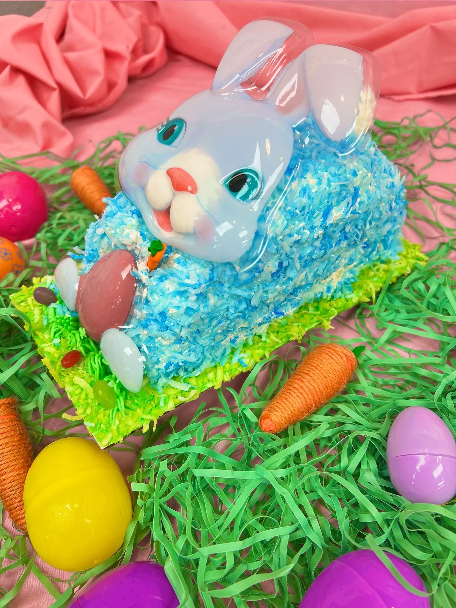 Easter bunny cake with blue coconut, candy face, carrots, plastic eggs, and green grass.