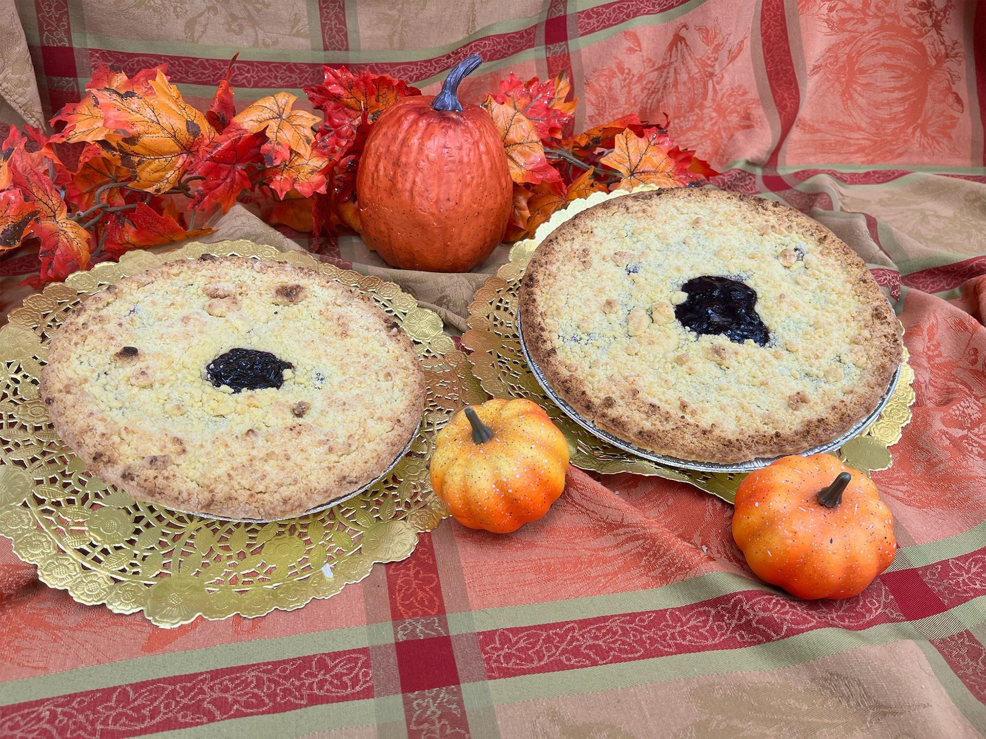 Two blueberry pies on gold doilies, pumpkins and fall leaves on plaid fabric.