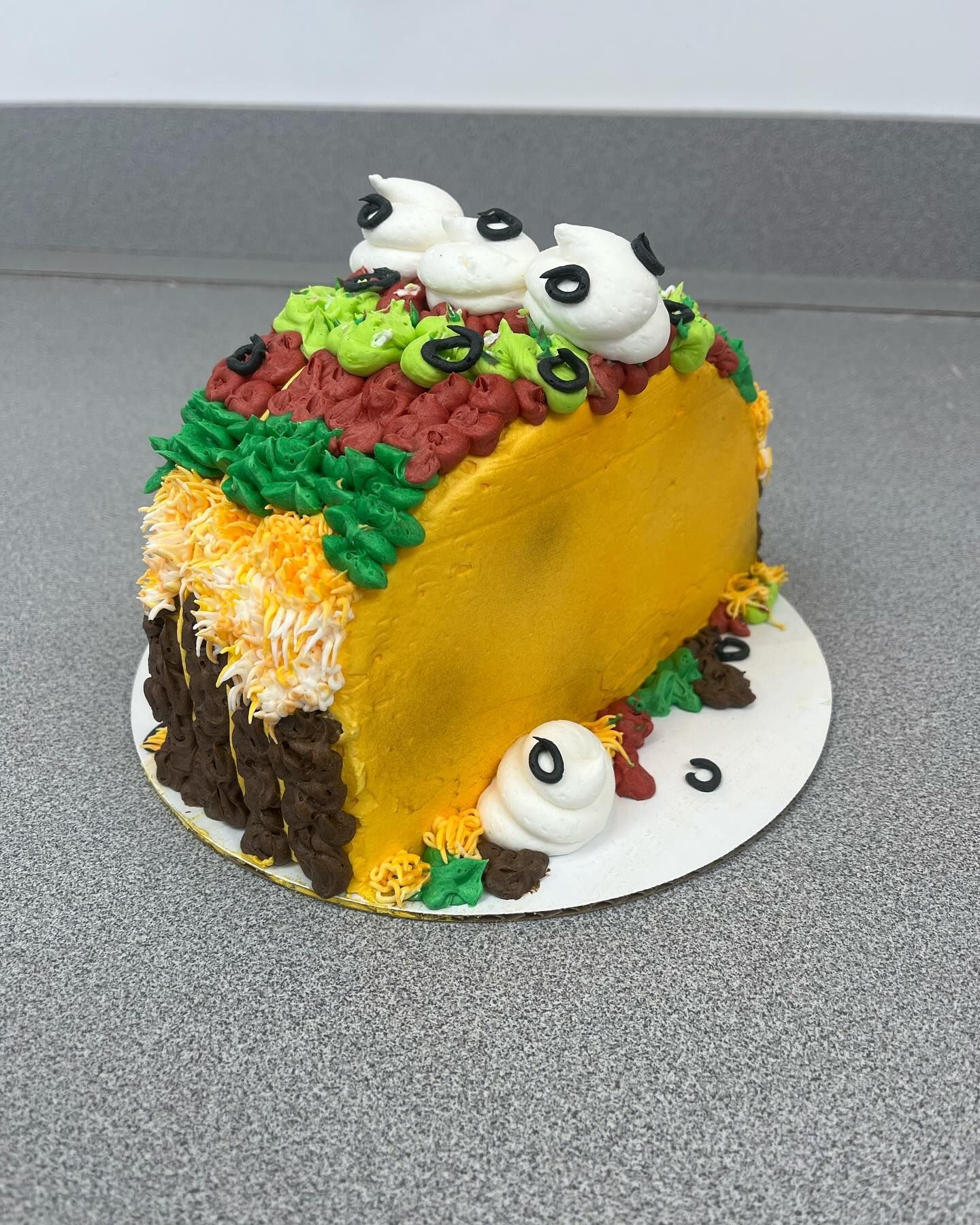 Taco-shaped cake on white plate; yellow shell, layered frosting: brown, orange, green, red, topped with cream swirls, and black olive decorations.