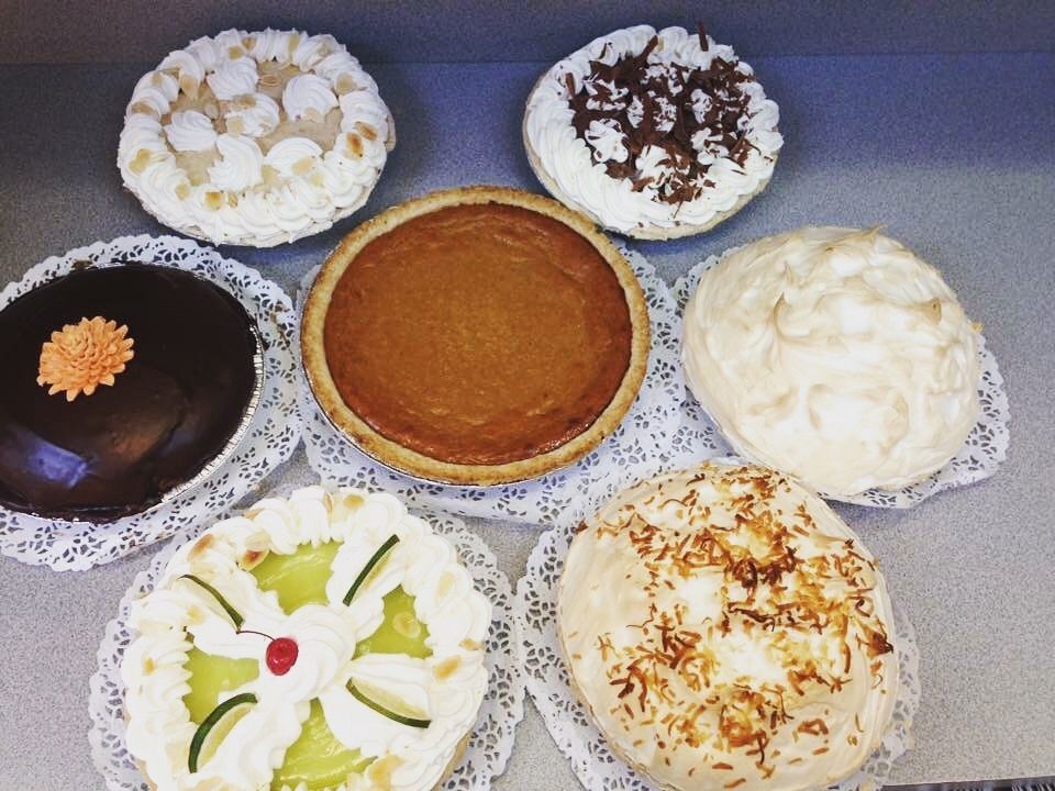 Assortment of seven decorated pies and cakes on doilies. Includes pumpkin pie and chocolate cake.