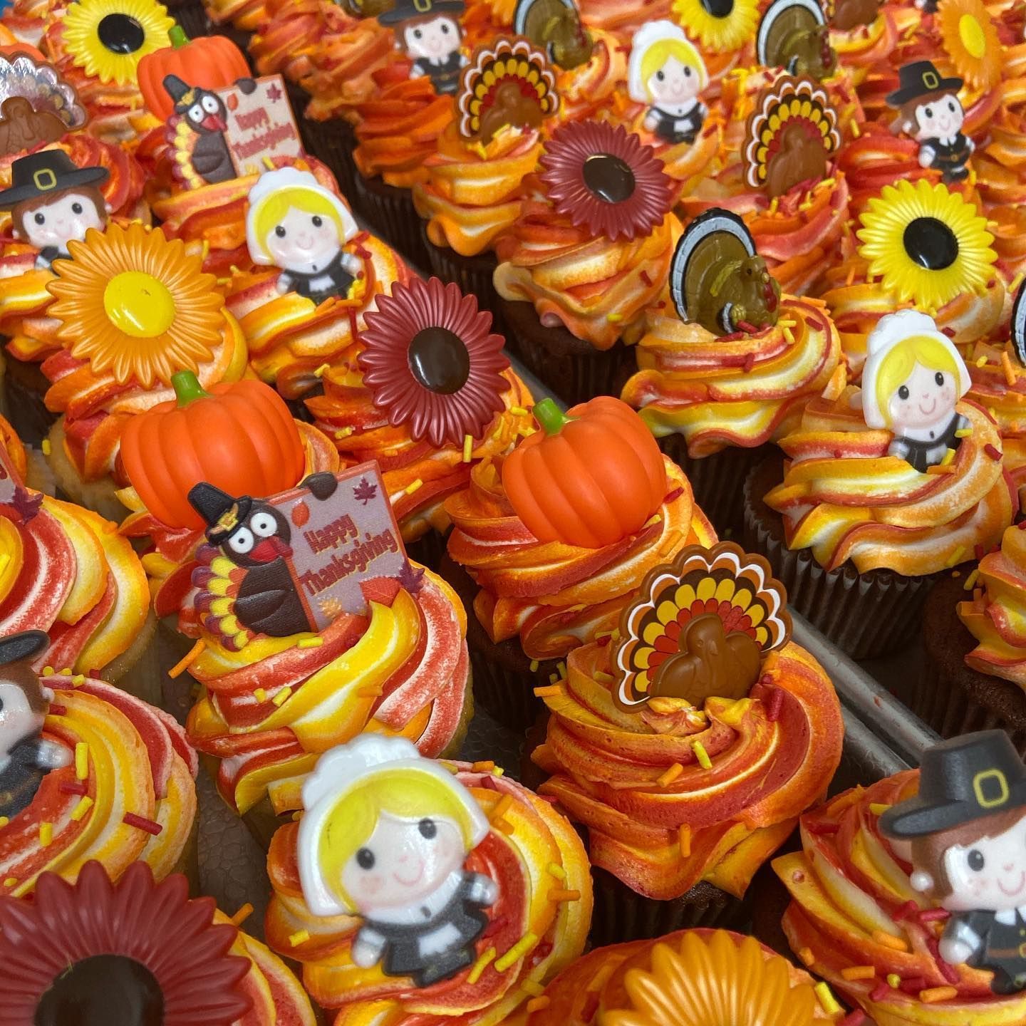 Thanksgiving cupcakes with orange and yellow frosting, topped with pumpkins, sunflowers, turkeys, and pilgrims.