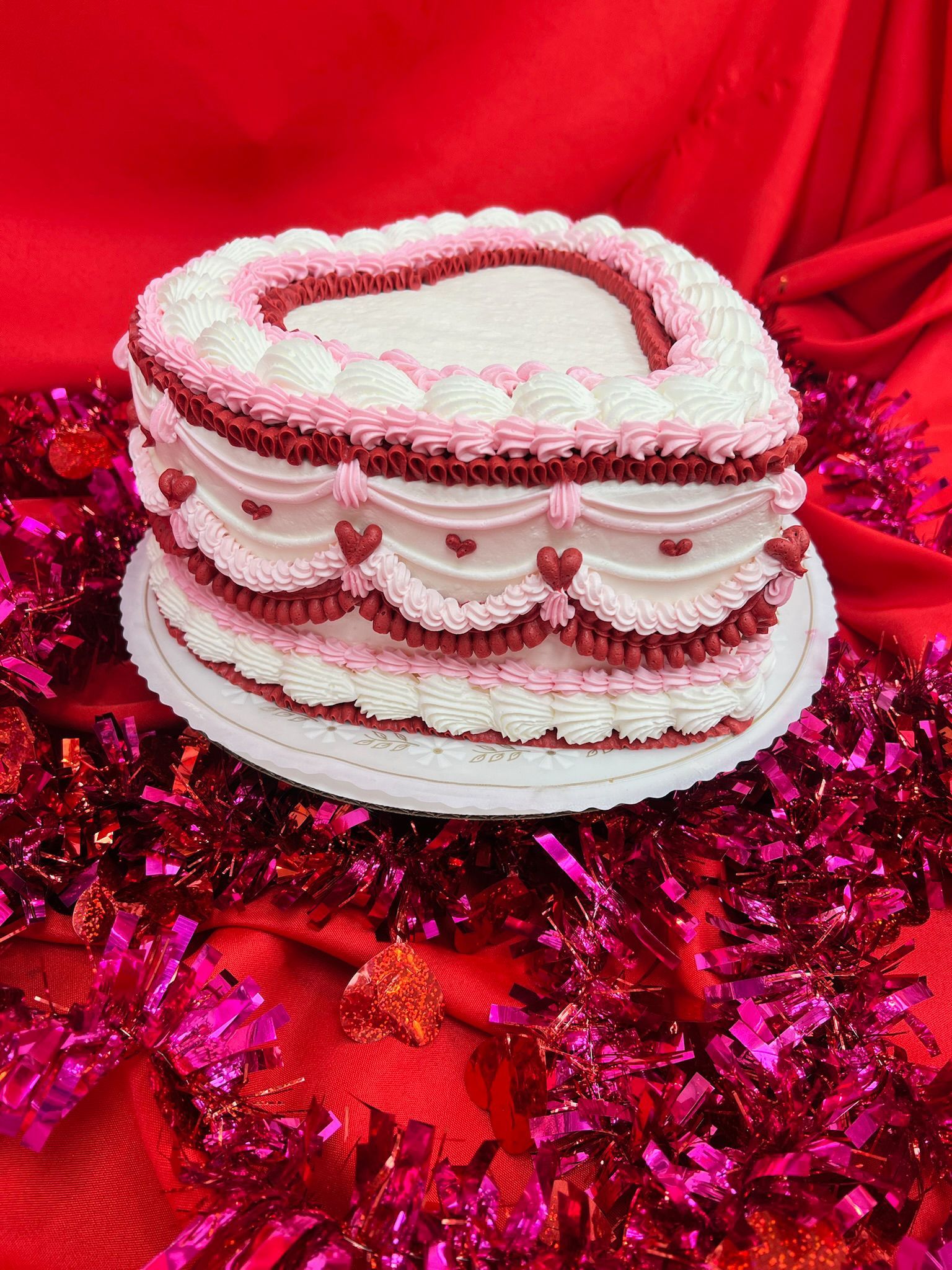 Heart-shaped cake with pink and white frosting, decorated on a white plate, on a red cloth with pink tinsel.