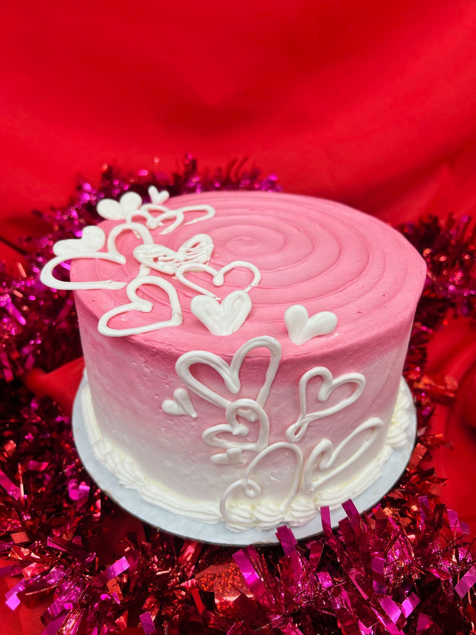 Pink ombre cake with white icing hearts on a red backdrop and pink tinsel.